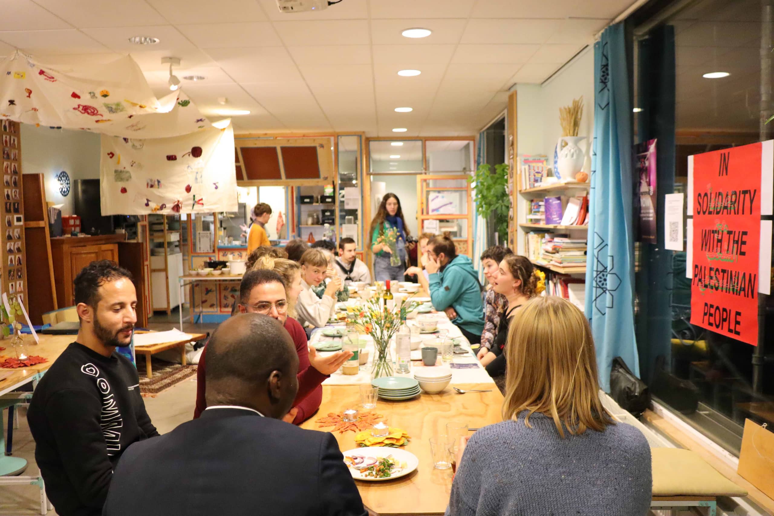 A long table of guests sharing a Community Dinner in the main room.