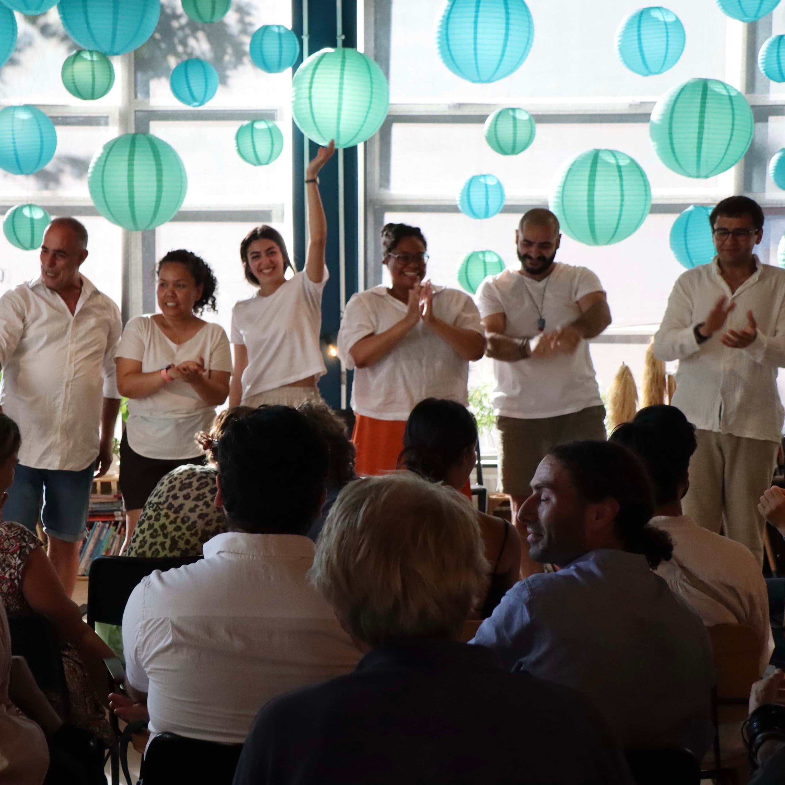 Performers in white shirts on stage in front of hanging paper lanterns, with the audience watching.