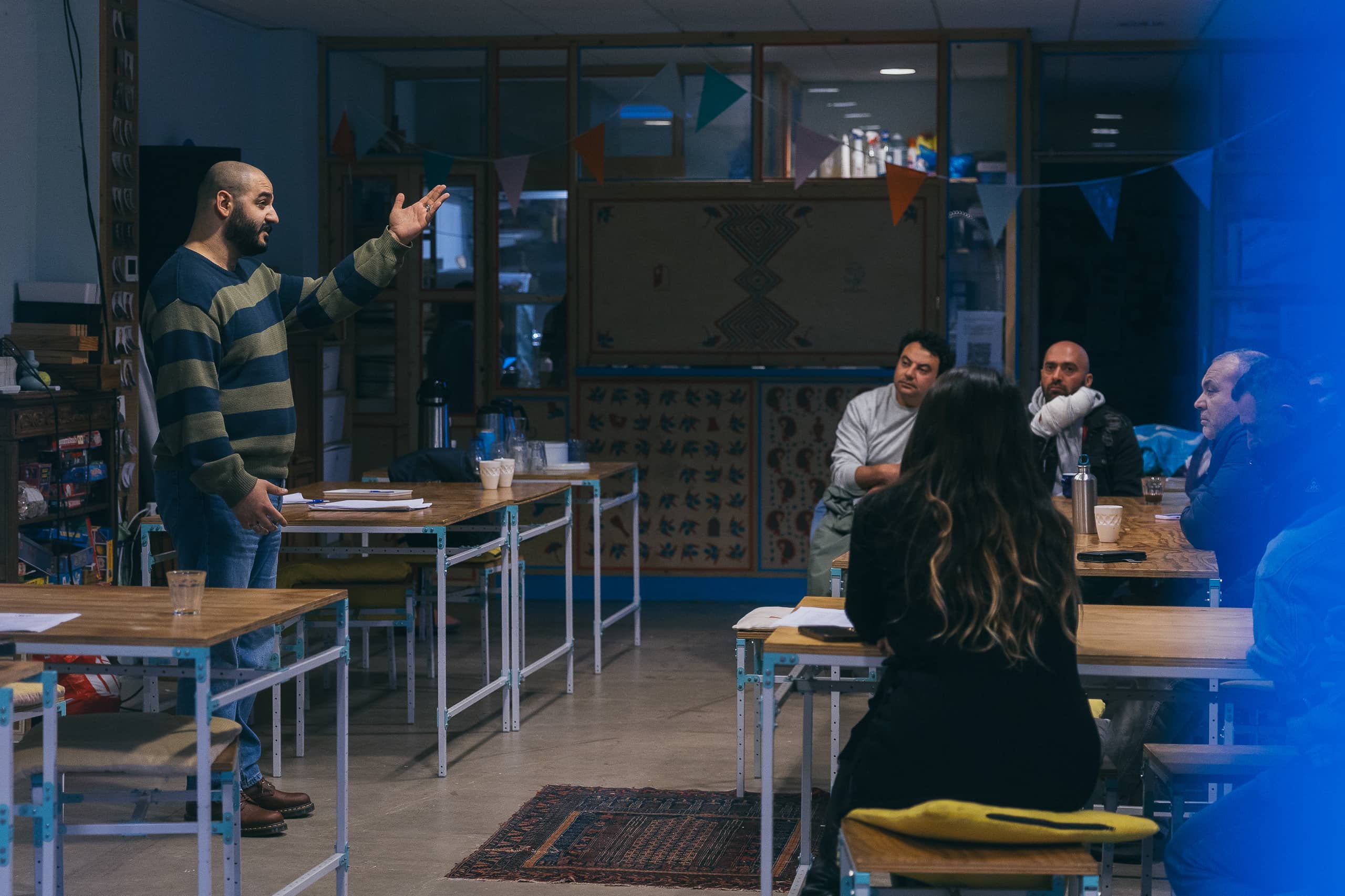 A photo of the storytelling course giver, at De Voorkamer, speaking in from of people (the participants of the workshop) as they are sitting on the tables.